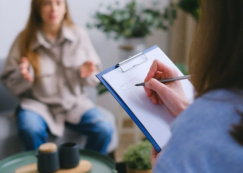 Unrecognizable female psychologist taking notes on clipboard while listening to patient sitting on blurred background during psychotherapy consultation in office