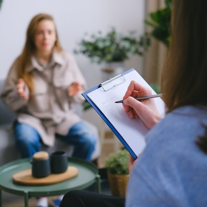 Unrecognizable female psychologist taking notes on clipboard while listening to patient sitting on blurred background during psychotherapy consultation in office